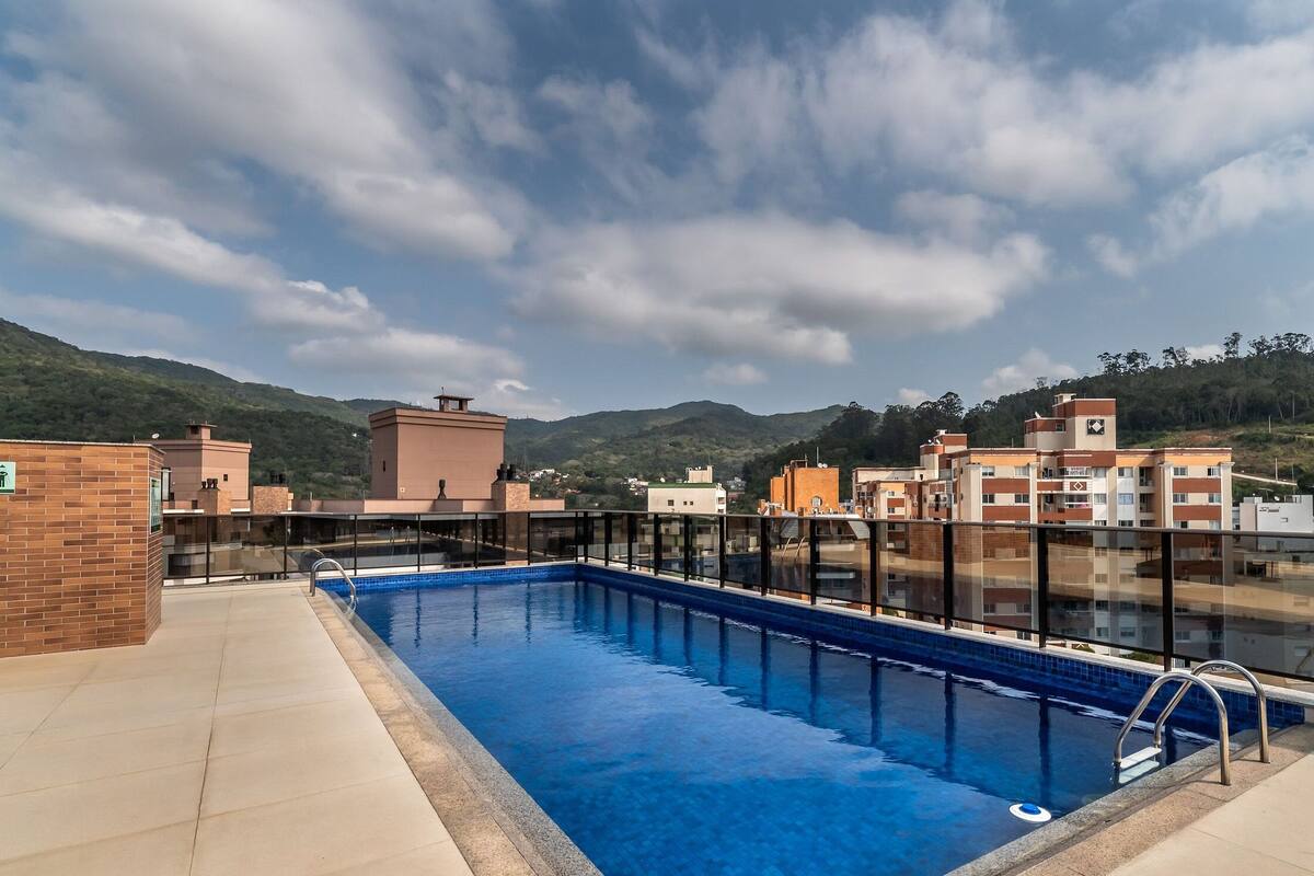 A rooftop swimming pool with a glass barrier, surrounded by buildings and a mountainous backdrop.