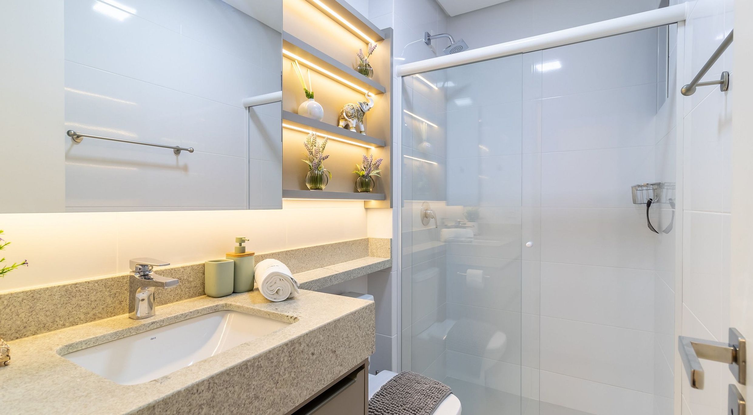Une salle de bain moderne avec une douche à parois de verre, un lavabo sur un plan de travail en granit et des vases décoratifs disposés sur une étagère.