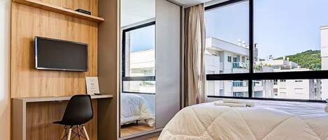 A hotel room with a bed, a desk with a chair, a TV, and a large window with a view of buildings.