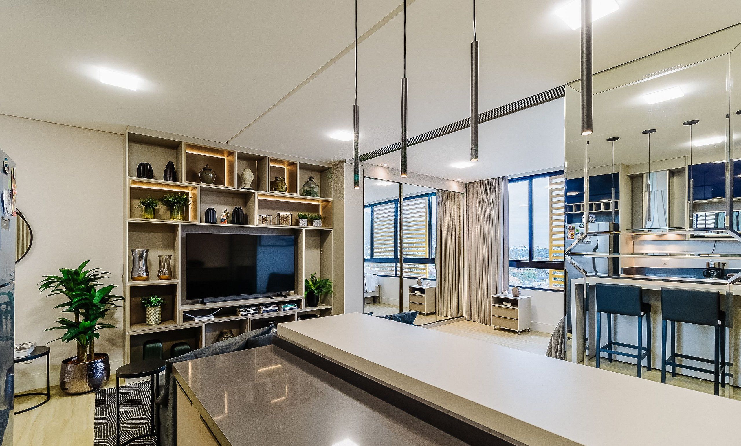 A modern living room with a large flat-screen TV, built-in shelves with decorative items, a dining area with a bar, and a large window with curtains.