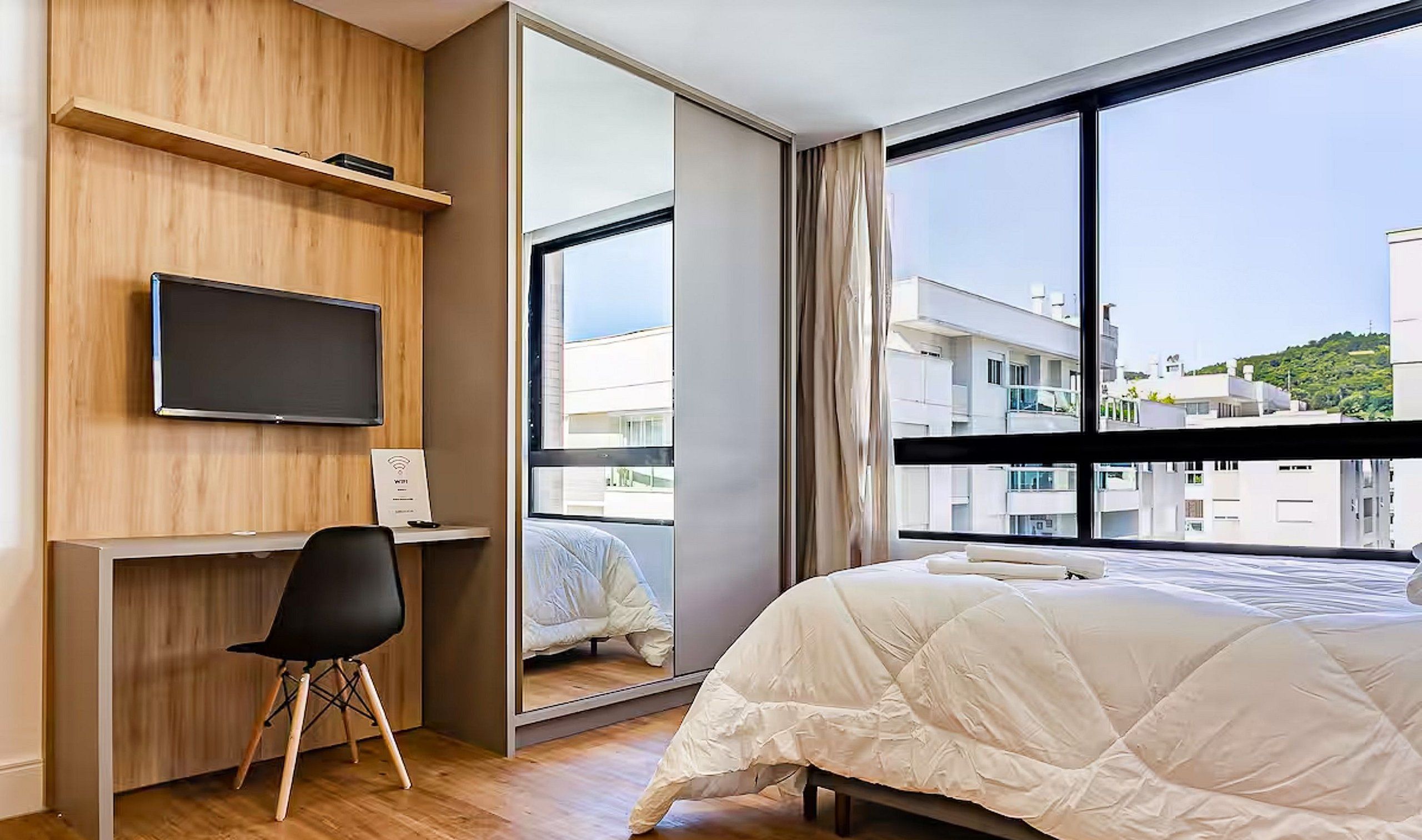 A hotel room with a bed, a desk with a chair, a television, and a large window offering a view of buildings and a hill.