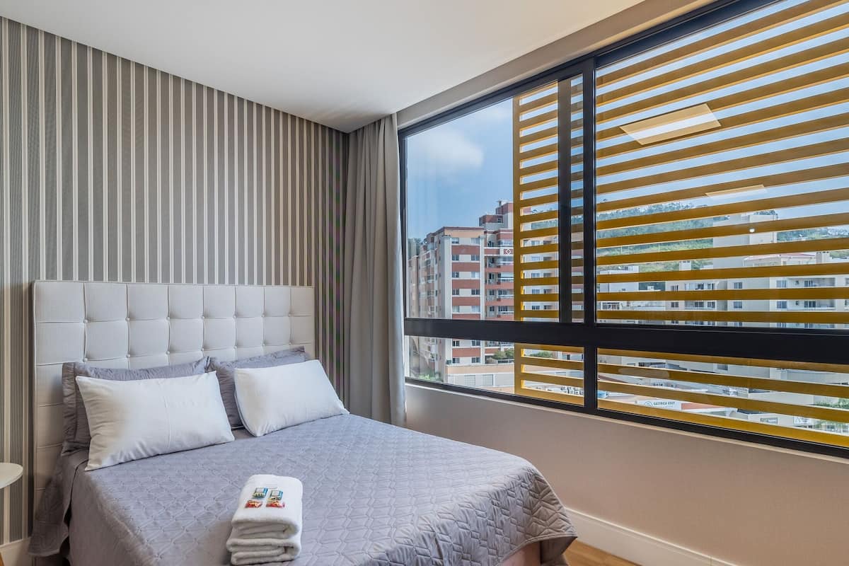 A hotel room with a large window, striped curtains, a tufted headboard, and a neatly made bed.