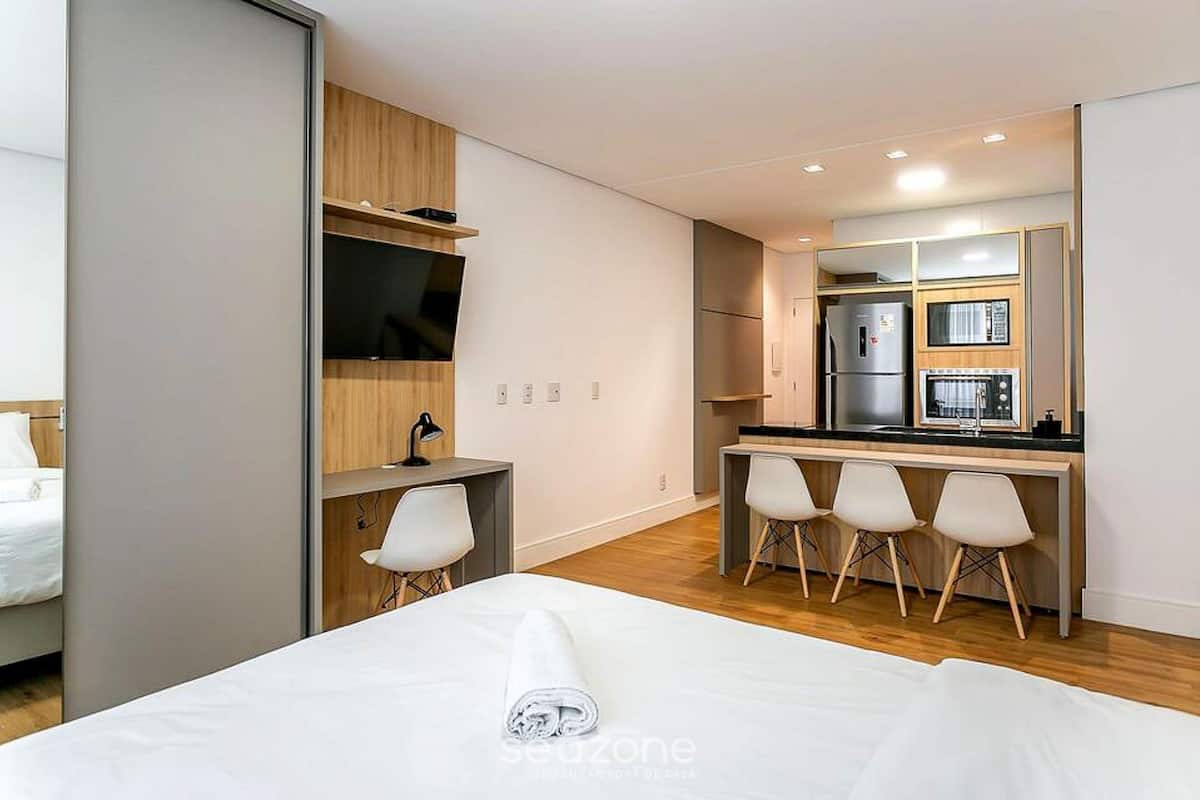 A modern hotel room with a bed, a desk, a TV, a small kitchenette, and a seating area with chairs.