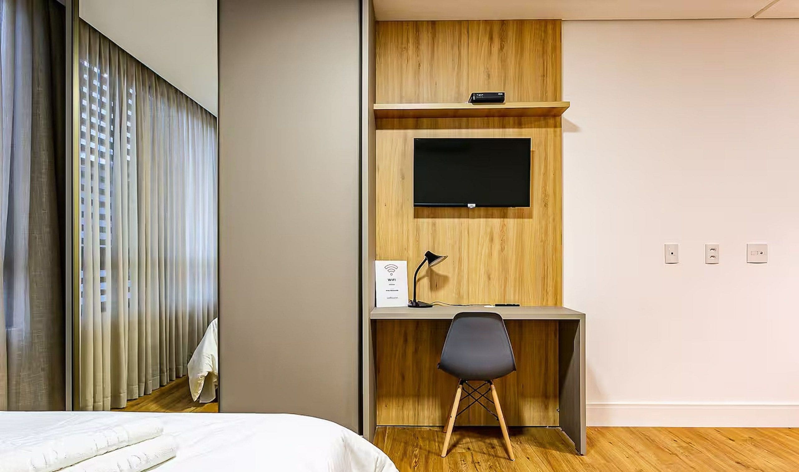 Una camera d'albergo con un letto, una scrivania con una sedia, una TV a schermo piatto, una lampada e una presa di corrente.