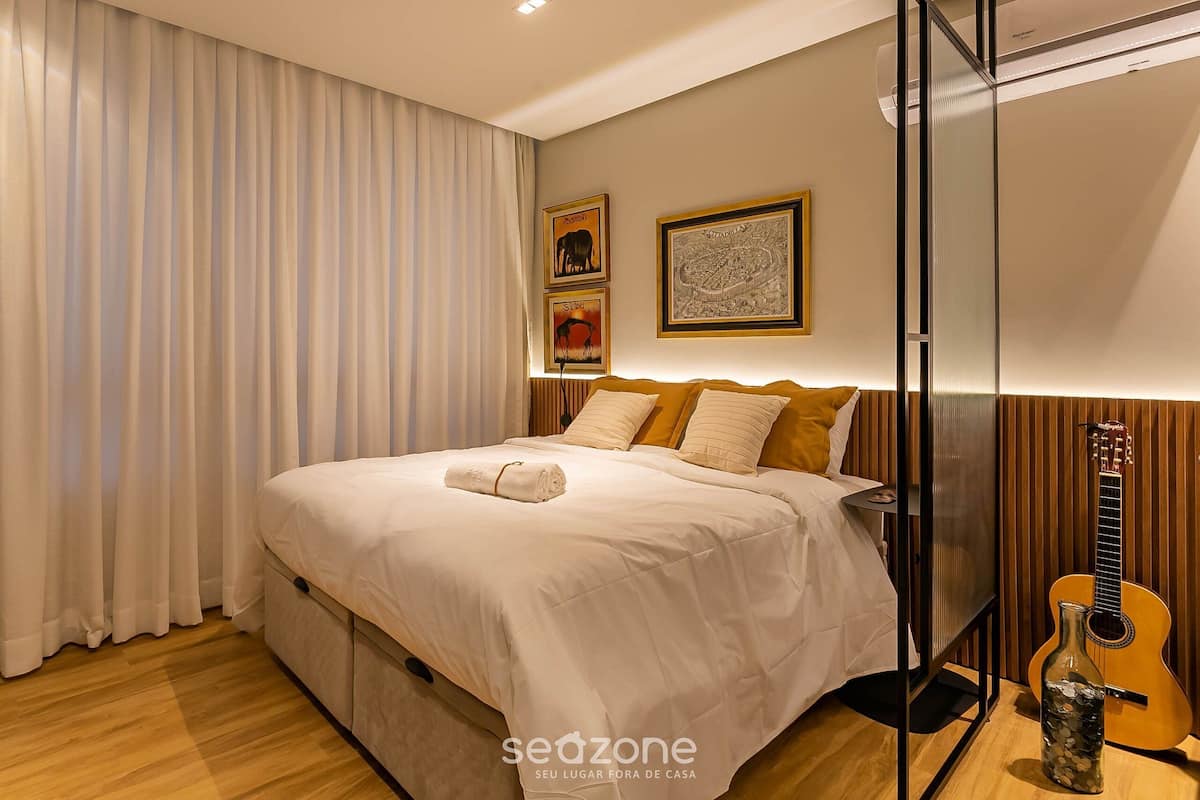 A neatly made bed with a rolled towel, a guitar, and a bottle of water on the floor.
