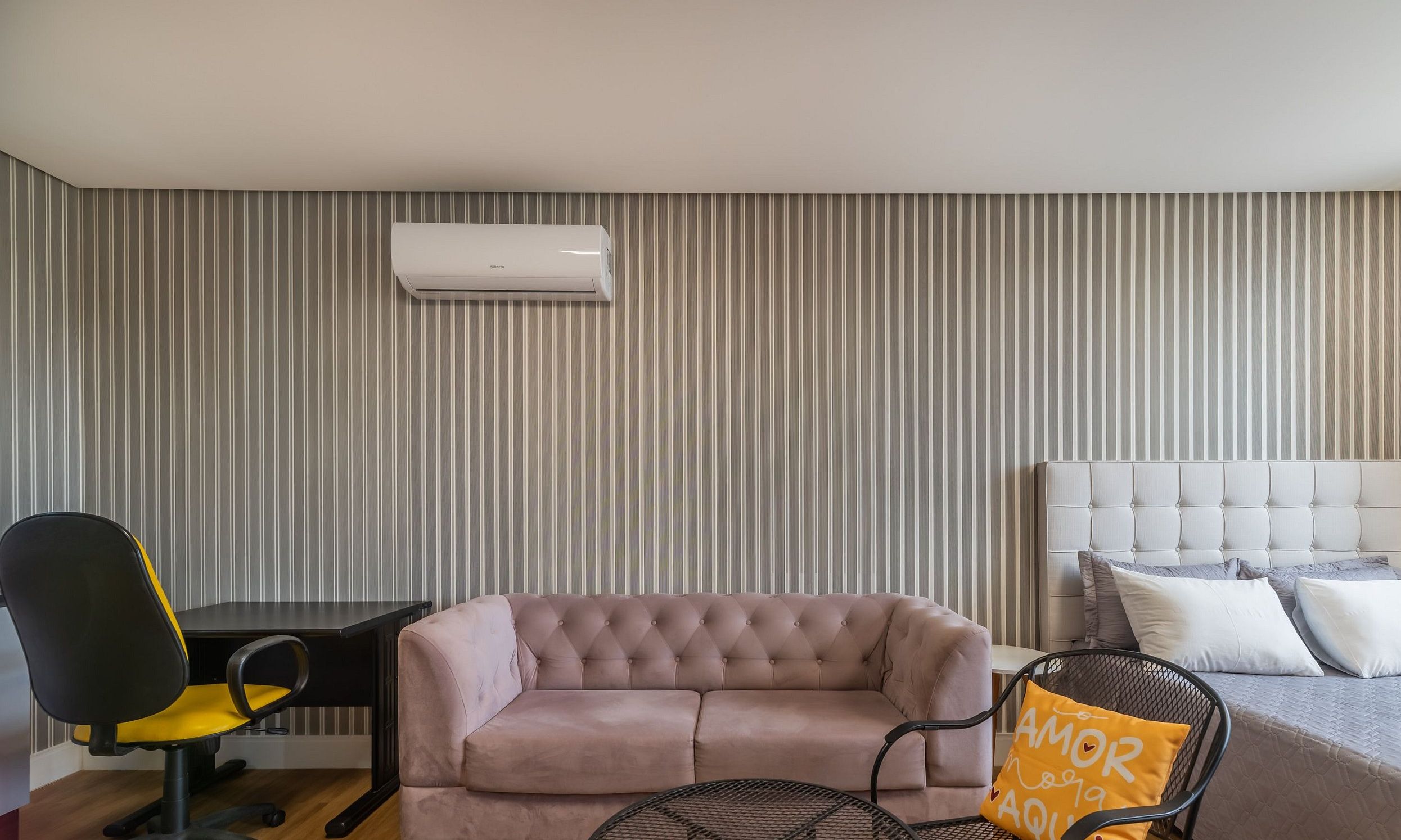 A room with a striped wall, a tufted sofa, a bed with a headboard, a desk with a chair, and an air conditioning unit.