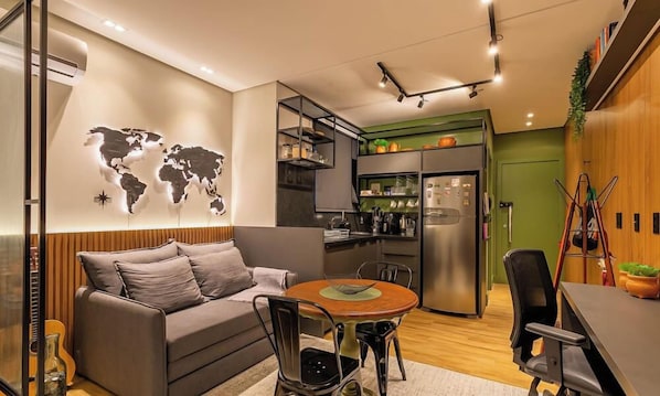 A modern living room with a sofa, a dining table, and chairs. There is a kitchen area with a refrigerator and shelves. - SSG Estúdios próximos a UFSC em Florianópolis (Florianópolis)
