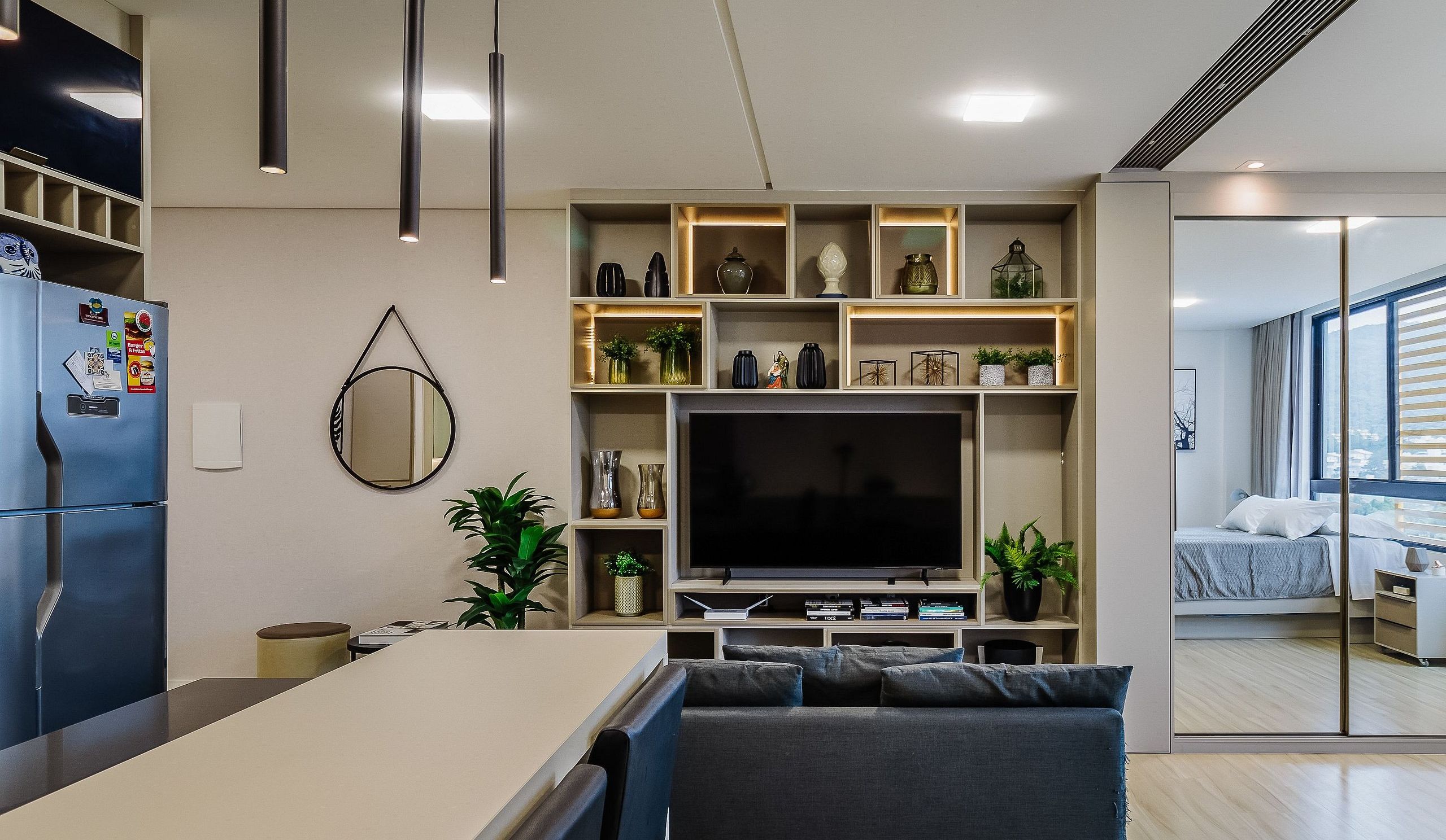 A modern kitchen with a central island, built-in shelving with decorative items, a large flat-screen TV, and a dining area with a grey sofa.