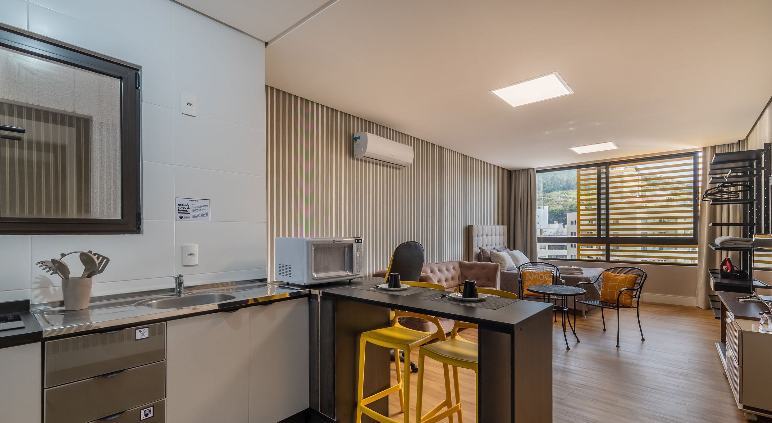 A modern kitchen with a microwave, sink, and bar stools. There is a dining area with a table and chairs, and a large window providing a view of the outside.