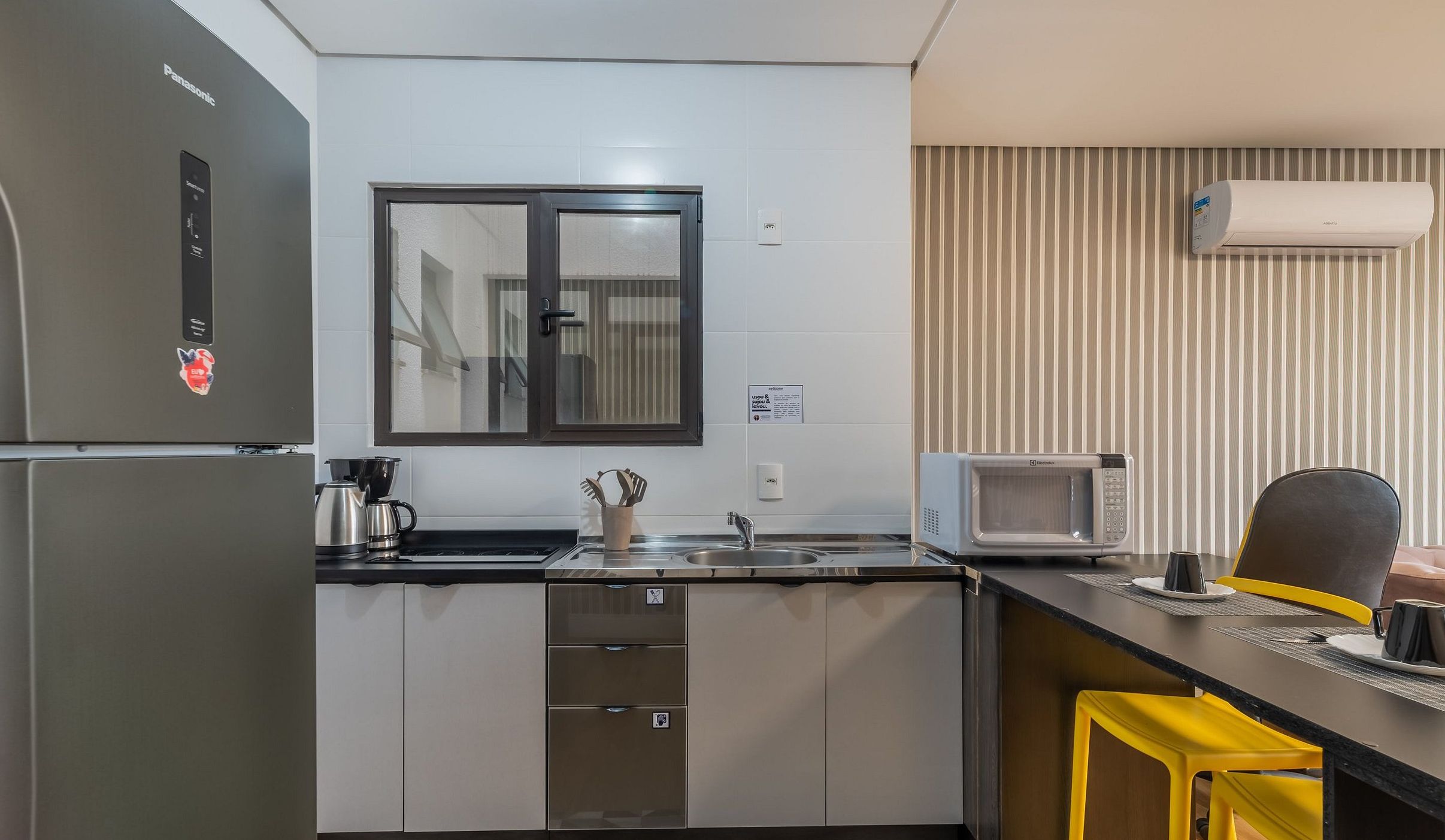 A compact kitchen with a refrigerator, microwave, sink, and a small dining area with a chair.