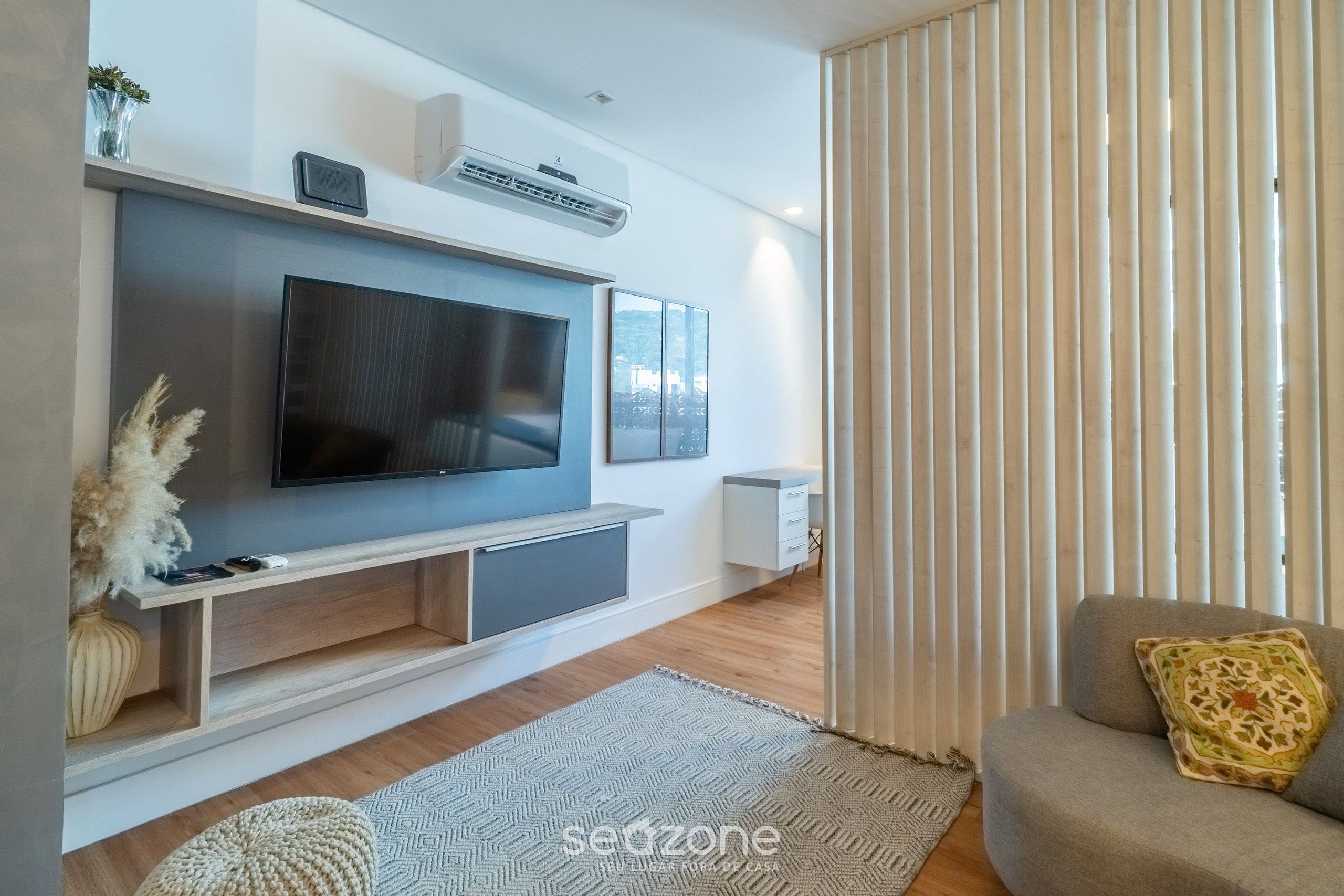 A modern living room with a flat-screen TV mounted on a wooden console, a white side table, and a grey sofa with a patterned cushion.