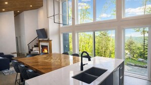 A modern kitchen with a large island, a fireplace, and large windows offering a view of the outdoors. - Havre du Mont * New Construction (st-come)