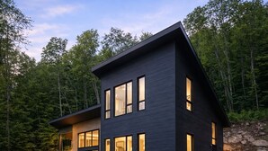 A modern two-story house with a dark exterior, large windows, and a well-maintained lawn. - Havre du Mont * New Construction (st-come)