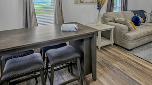 Un salon moderne avec une table à manger en bois, deux tabourets de bar, un canapé et un tableau accroché au mur.