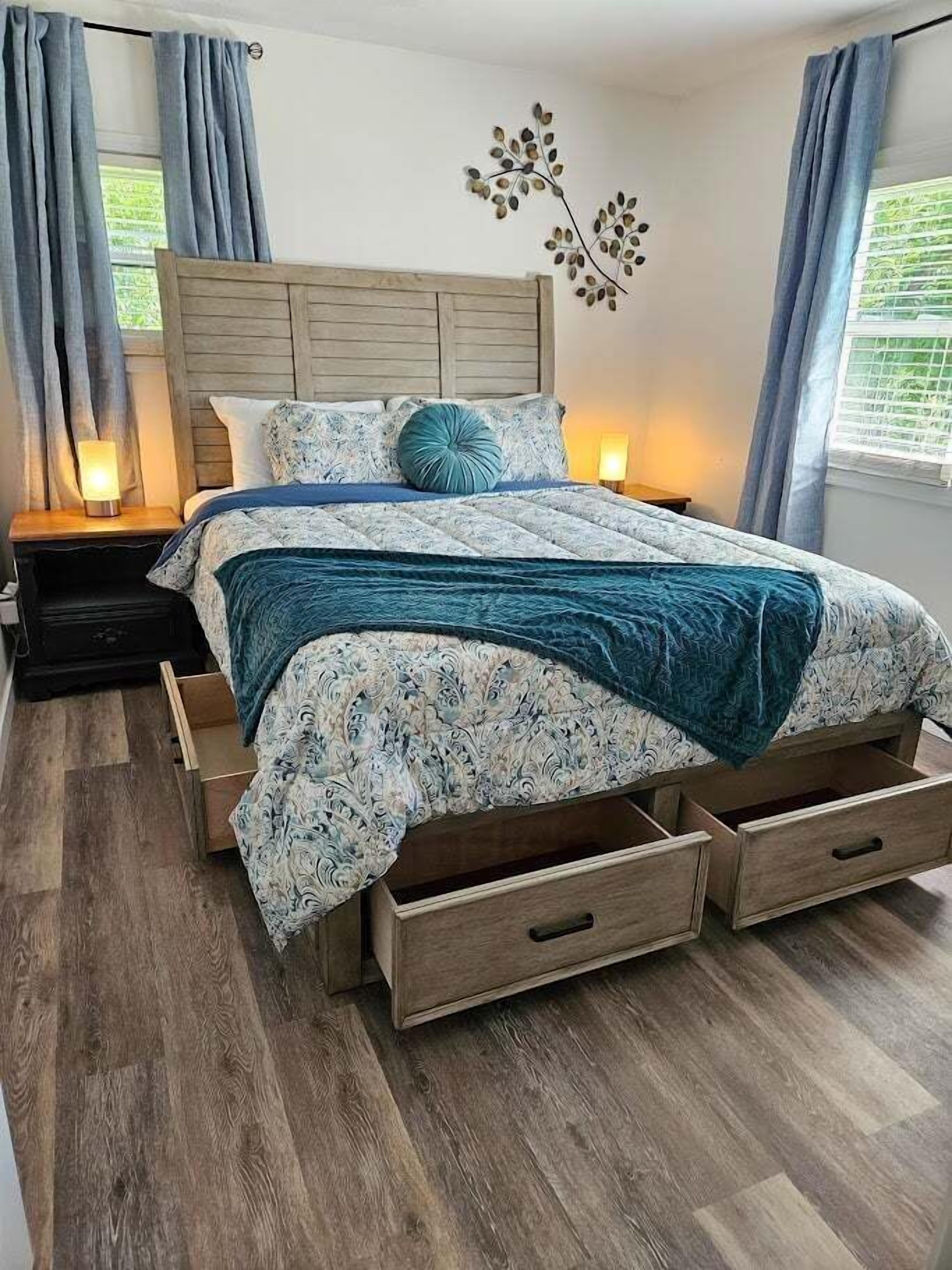 A bedroom with a wooden headboard, a bed with patterned bedding, two bedside tables with lamps, and a wall-mounted decorative piece.