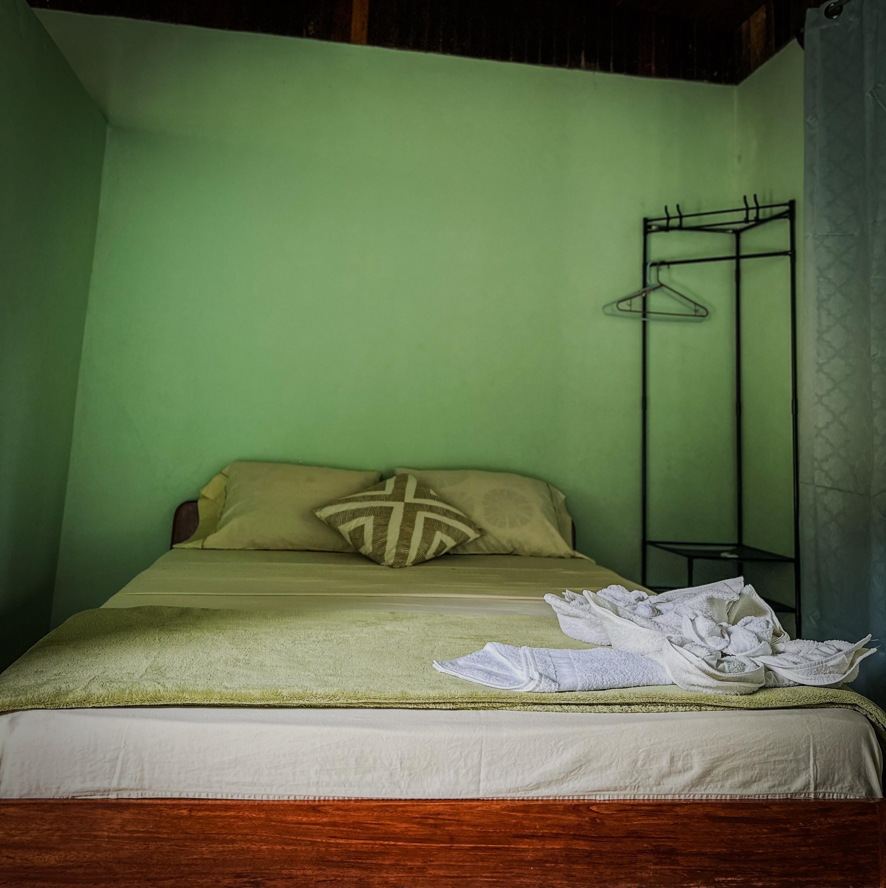 Ein Einzelbett mit einer grünen Wand, einem Holz-Kopfstück und einem Metallgestell mit Kleidung.