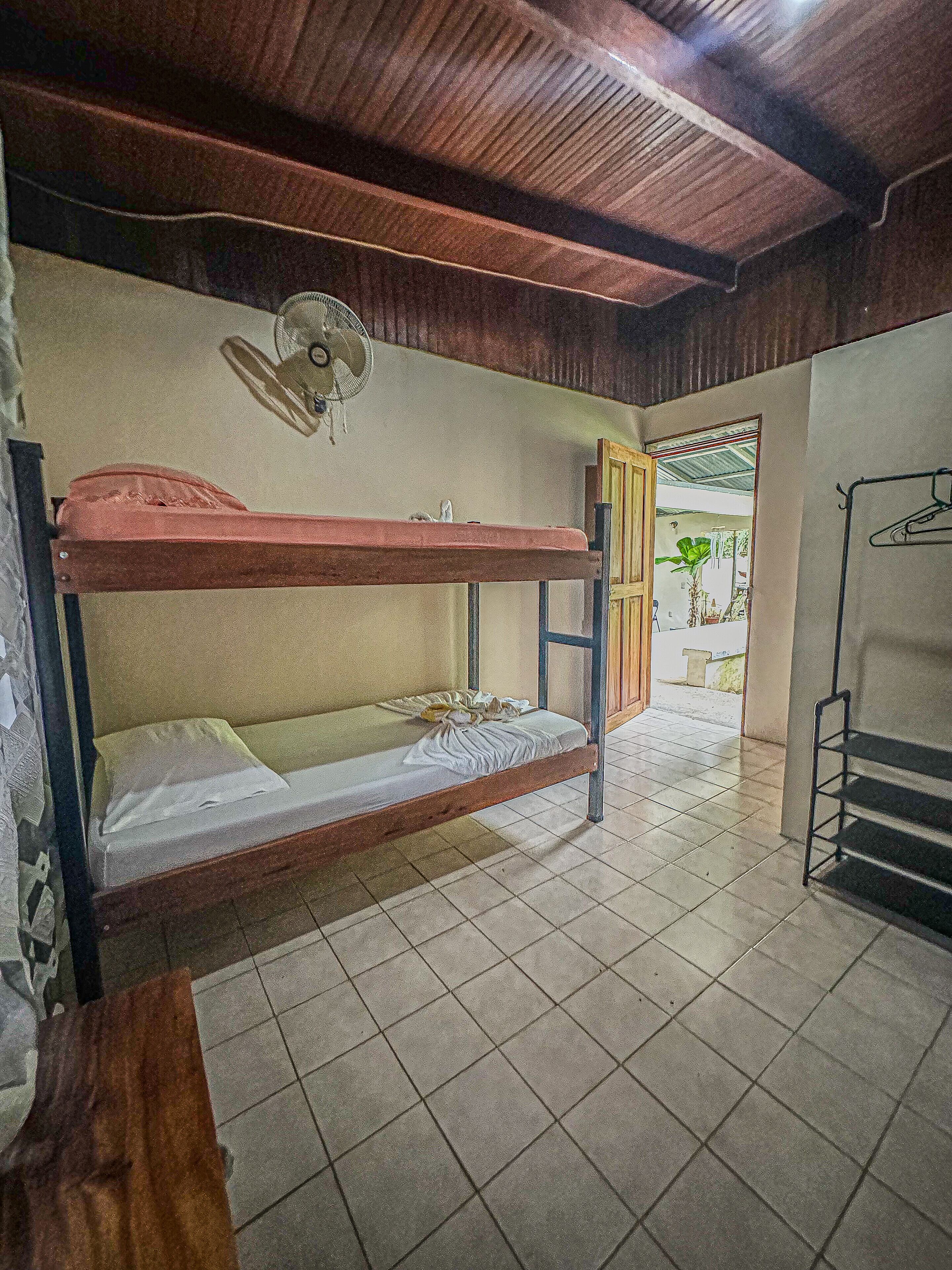 Een kamer met een stapelbed, een ventilator, betegelde vloer en een deur naar een buitenruimte.