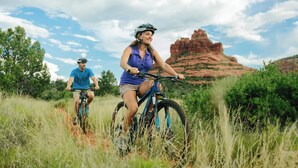 Dos personas montando en bicicleta en un campo cubierto de hierba, con una formación rocosa roja al fondo.