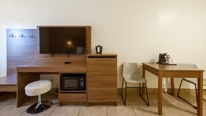 Una habitación compacta con escritorio de madera, televisión, microondas, un taburete pequeño, dos sillas y cafetera.