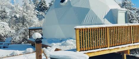 Una estructura en forma de cúpula cubierta de nieve con una terraza de madera y un banco.
