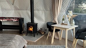 Een gezellig tent-achtig vertrek met een houtkachel, een bed, een tafel met een fles en glazen, en uitzicht op de natuur.