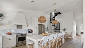 Una cocina moderna con armarios blancos, una isla central, sillas de mimbre y una escalera en espiral negra.
