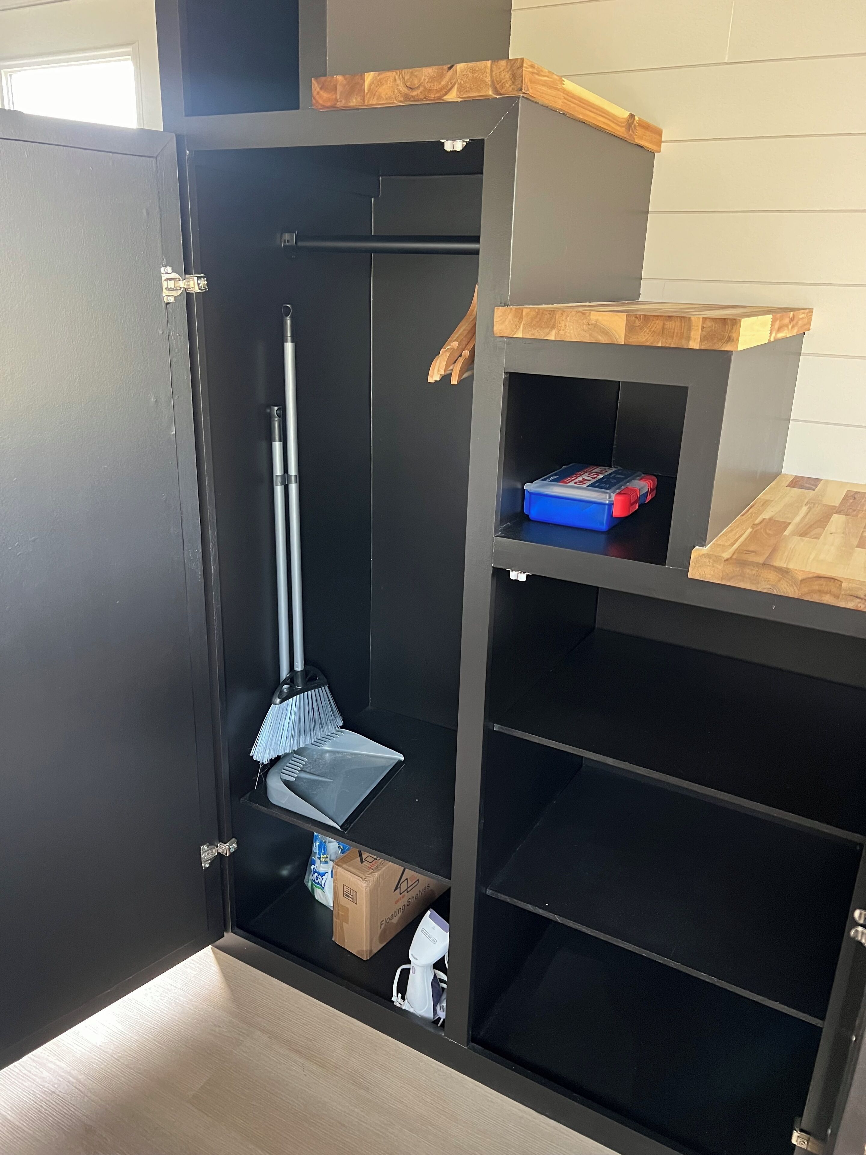 Una habitación con un armario negro, un escoba, un recogedor y un caja azul para guardar cosas.