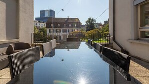 A sunlit outdoor dining area with a long table and wicker chairs, surrounded by buildings and a clear sky. - KAYA Zurich Stauffacher  (Zürich)