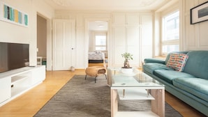 A modern living room with a flat-screen TV, a glass coffee table, a teal sofa, and a wicker chair. - KAYA Zurich Stauffacher  (Zürich)