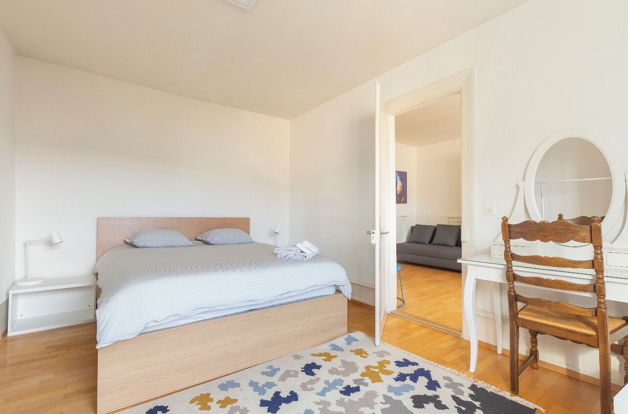 Un dormitorio con una cama, una mesita de noche, una silla, un espejo y una puerta que da a otra habitación.