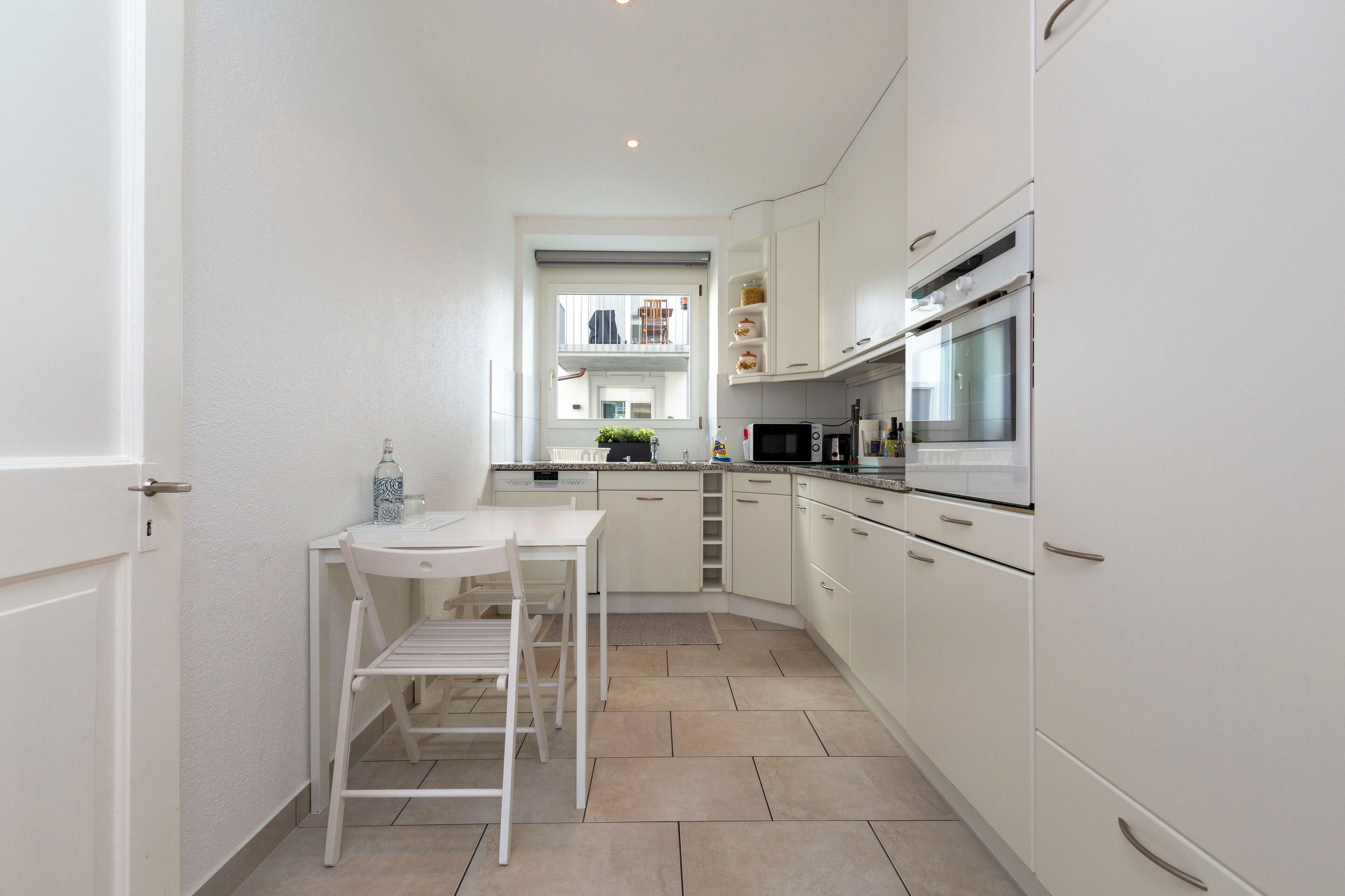 Cocina compacta con armarios blancos, microondas, horno y un pequeño comedor con mesa y silla.