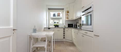 Cocina compacta con armarios blancos, microondas, horno y un pequeño comedor con mesa y silla.