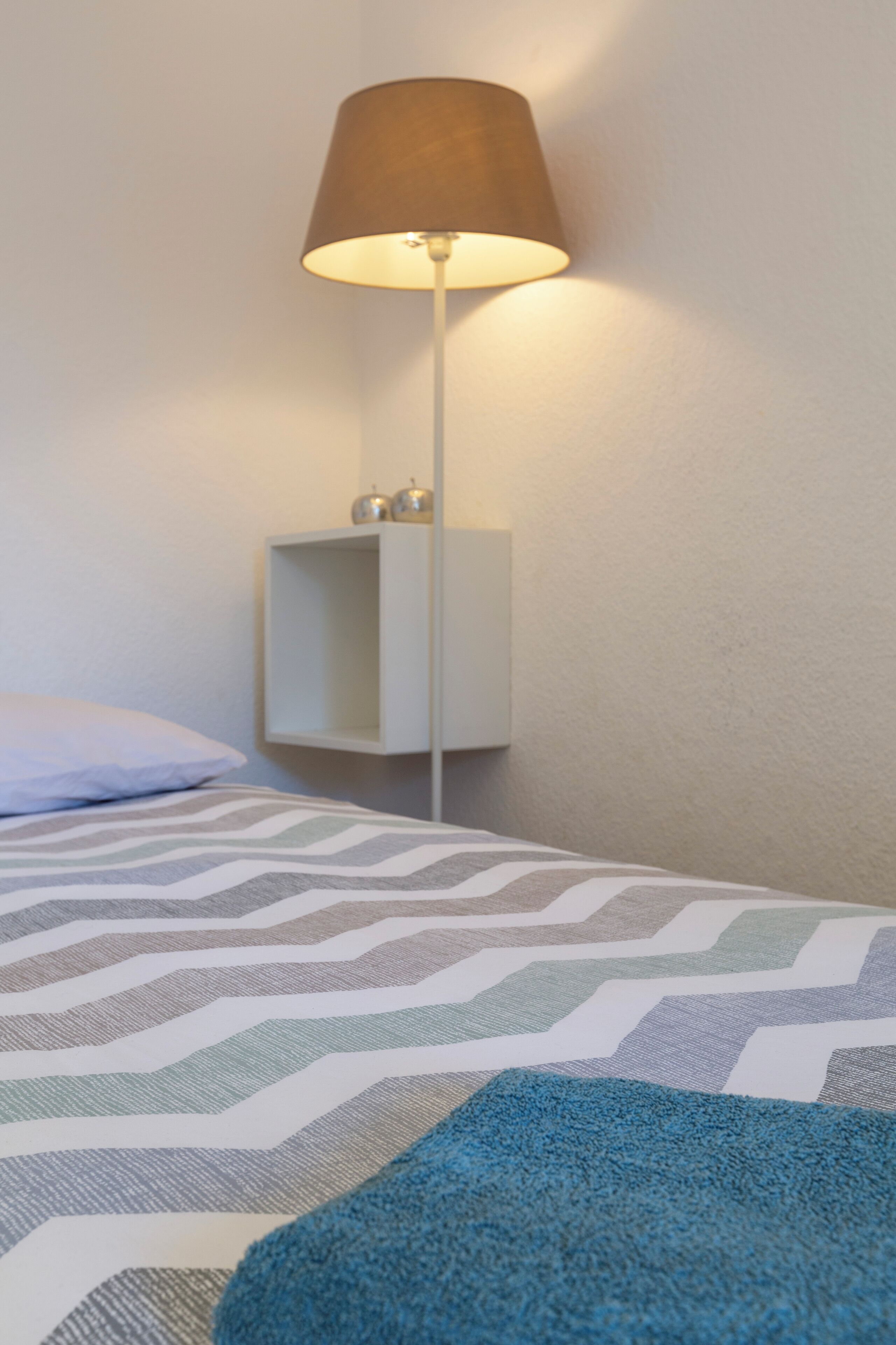 A neatly made bed with a patterned bedspread and a blue throw blanket. There is a white bedside table with a lamp and a small shelf.