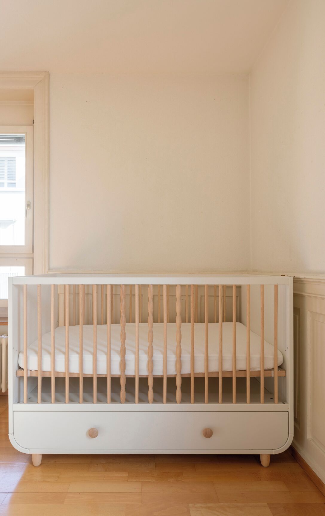 Una cuna blanca con listones de madera en una habitación con suelo de madera y paredes blancas.