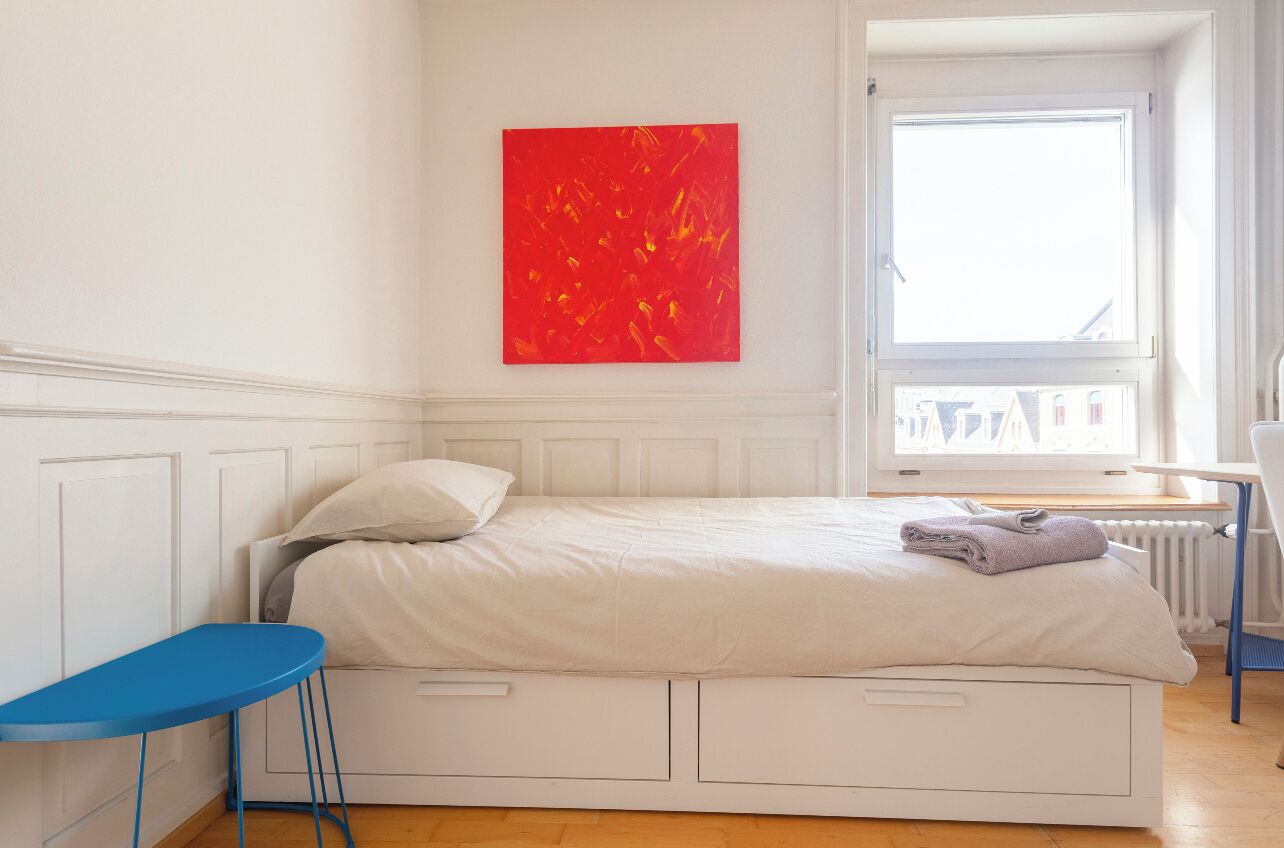 Une chambre à coucher avec un lit, une chaise bleue, un tableau abstrait rouge et une fenêtre donnant sur un paysage.