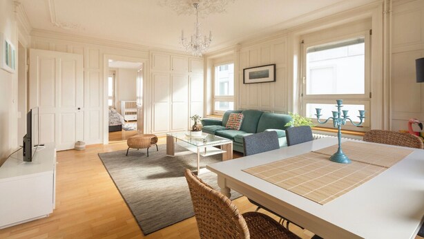 A spacious living room with a white ceiling, a chandelier, a sofa, a coffee table, and a dining table with a placemat. - KAYA Zurich Stauffacher  (Zürich)