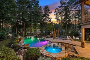 A swimming pool with a hot tub, surrounded by a stone deck and lounge chairs, set against a backdrop of tall trees and a sunset. - Livin' Large Lake Norman Estate w/ Pool, Dock, Game Room & Scenic Views (Mooresville)