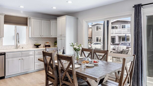 Una cocina moderna con armarios blancos, una mesa de comedor de madera y vistas a un barrio residencial a través de amplios ventanales.