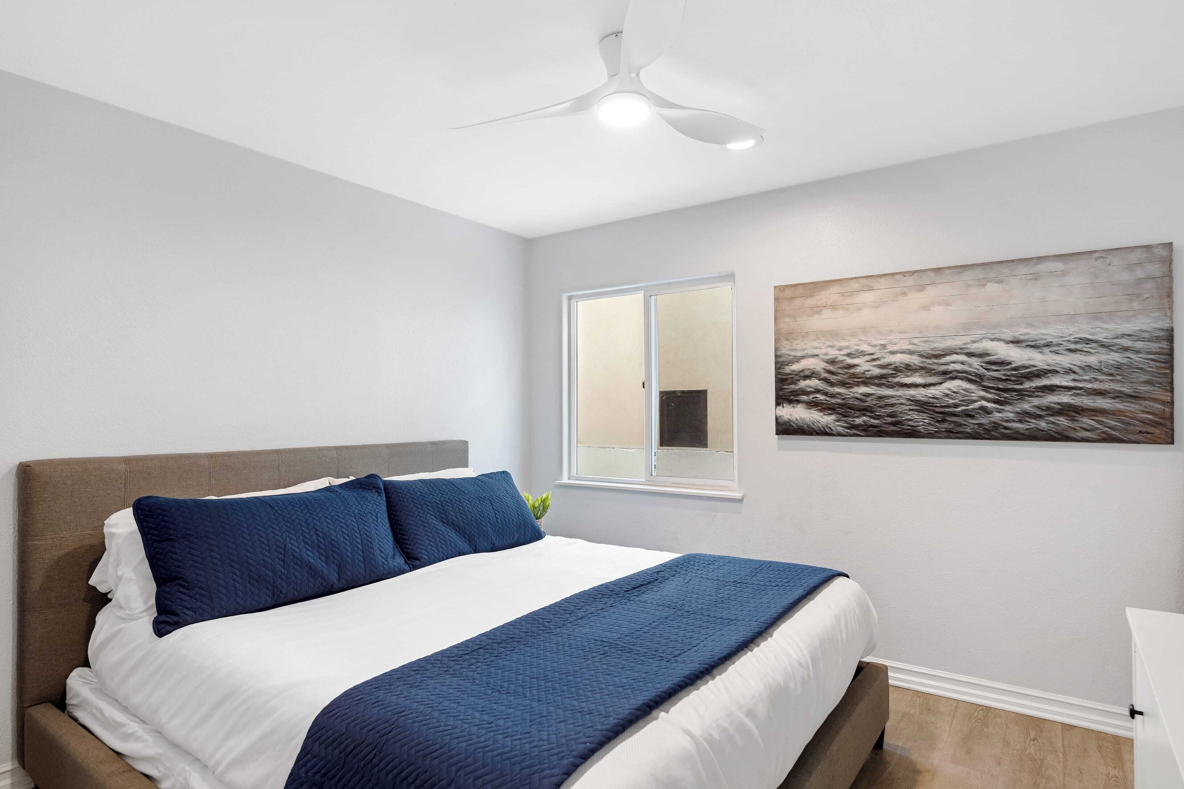 A bedroom with a bed, a ceiling fan, a window, and a painting on the wall.