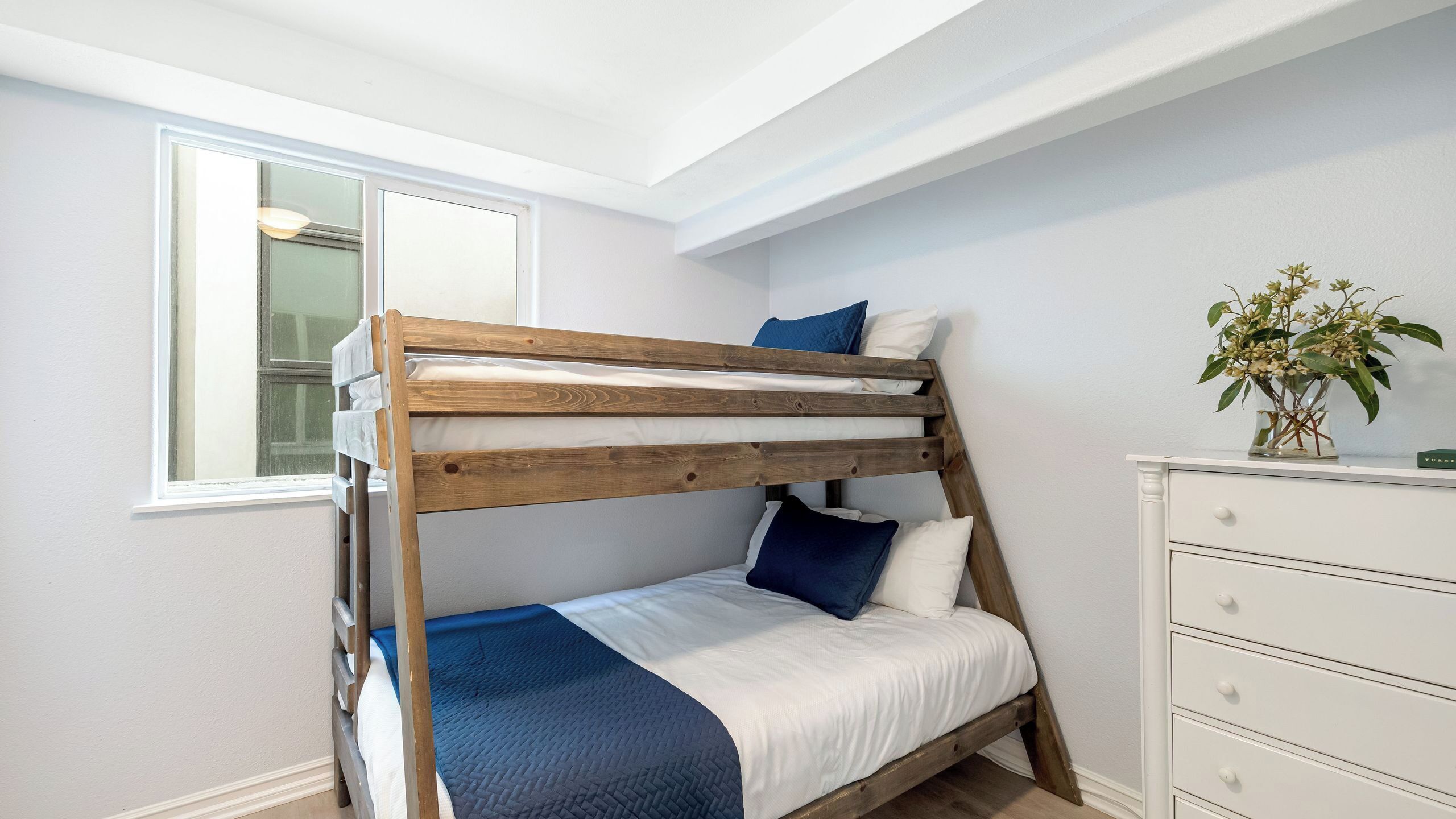 A bunk bed with a wooden frame, white bedding, and blue accents. A window with blinds is visible, and a plant is on a dresser.