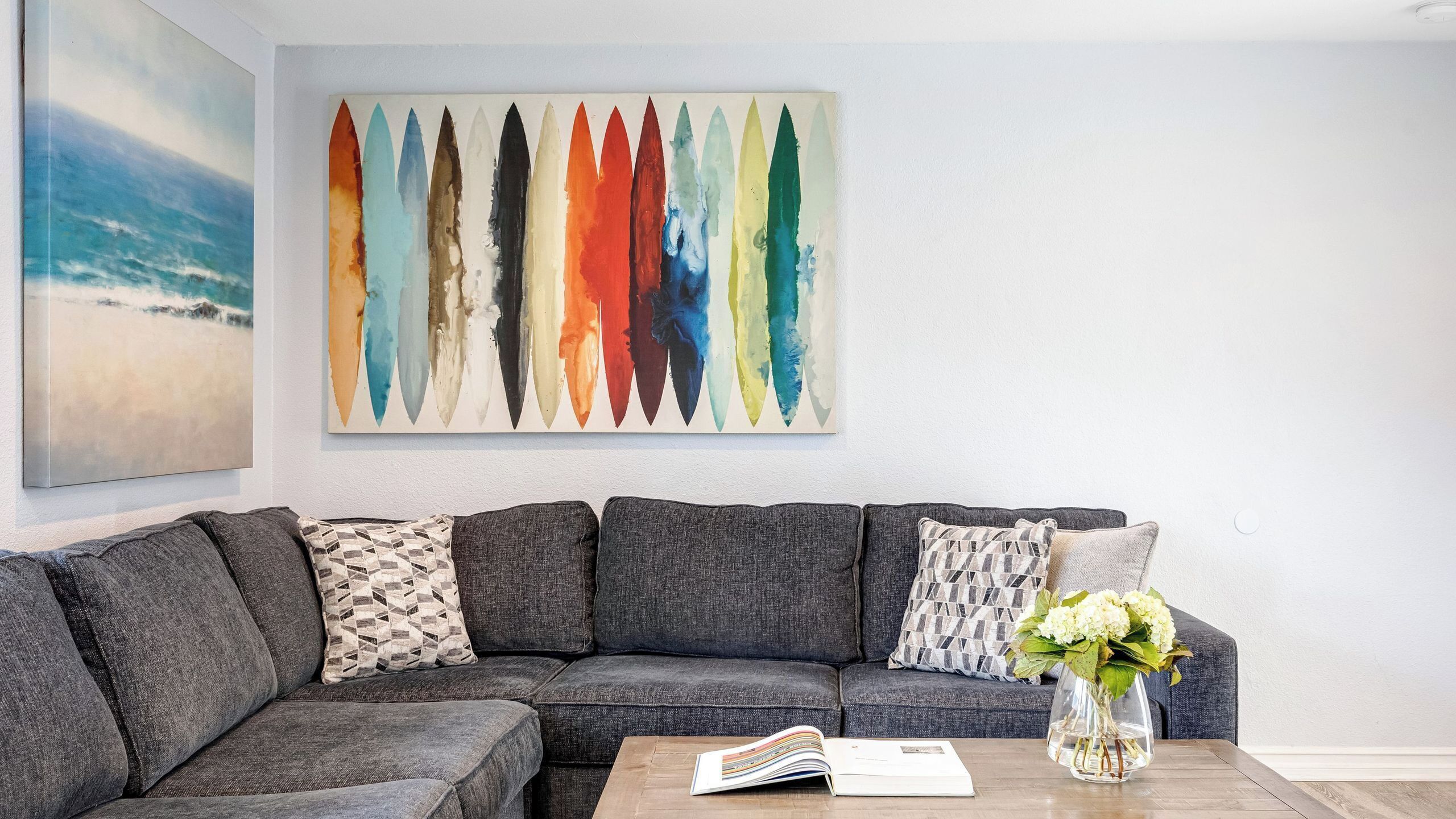 A grey sectional sofa with patterned cushions, a coffee table with a book, and a colorful abstract painting above.