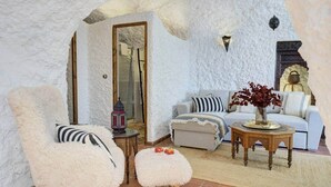 A cozy living room with a white textured wall, a wooden-framed mirror, a sofa with striped pillows, a small round table with a lantern, and a fluffy white pouf.