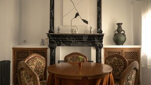A dining room with a round wooden table, floral upholstered chairs, a black marble fireplace, and a large vase on the mantel.