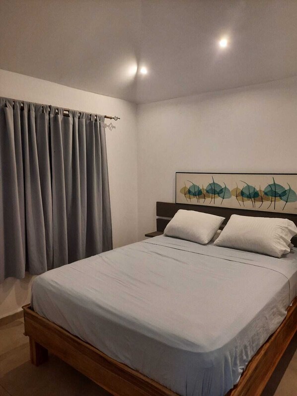A neatly made bed with a light-colored bedspread and two pillows, a wooden headboard, and a wall with a leaf-patterned artwork.