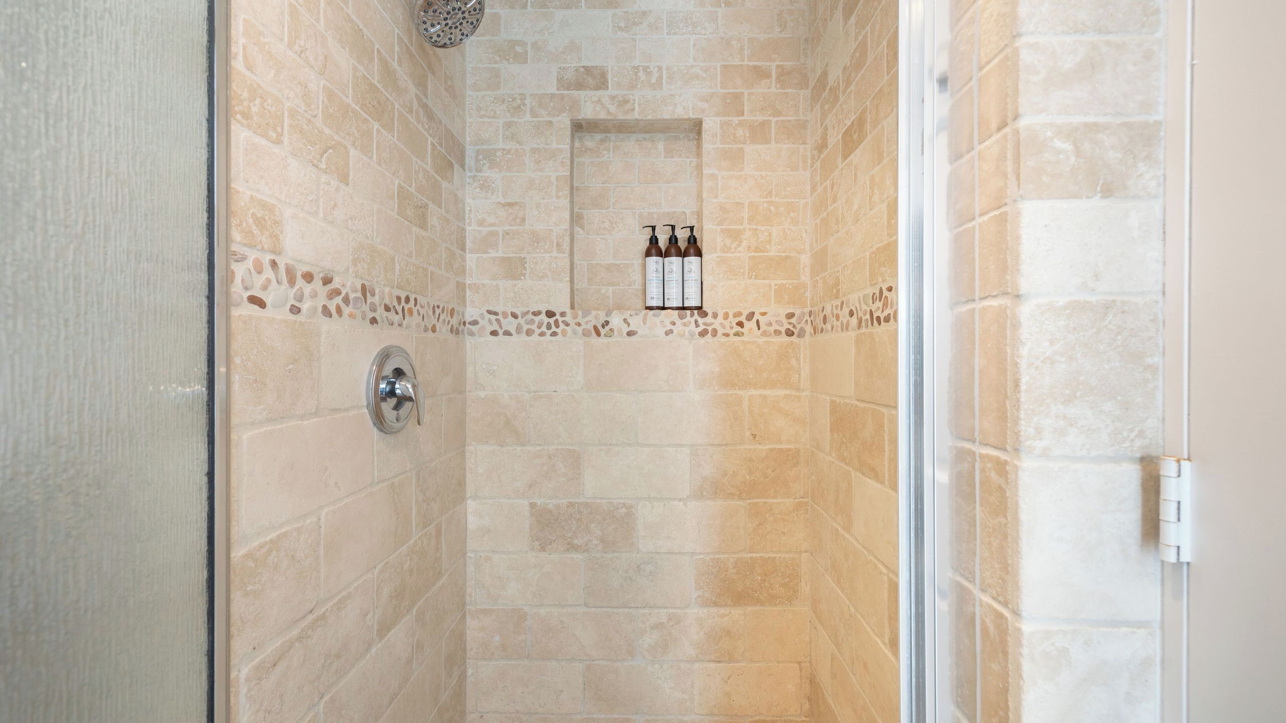 A shower with beige tiles, a glass door, and a shelf with three bottles.