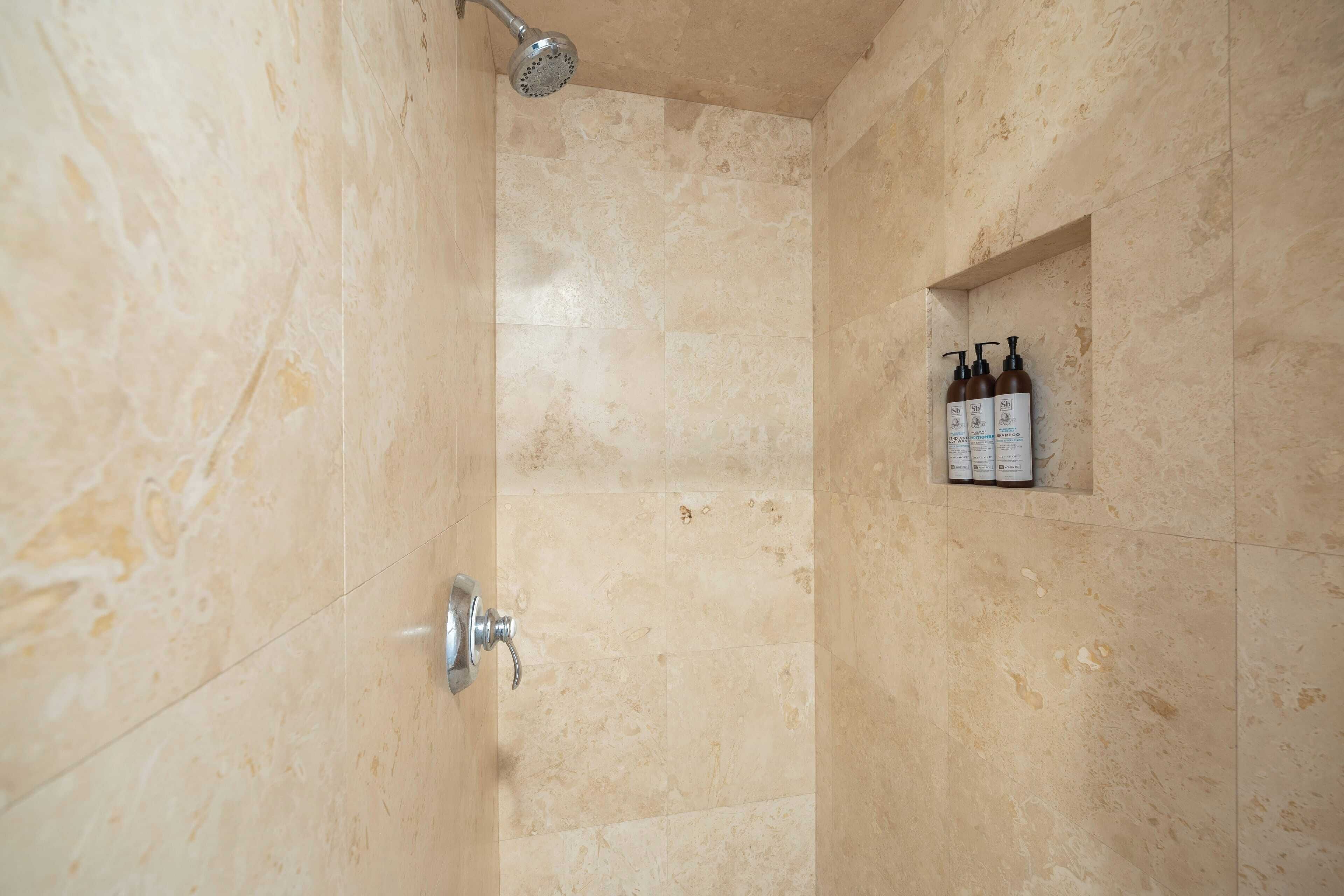 A shower with beige tiles and a handheld showerhead. There are three bottles of shampoo on a shelf.