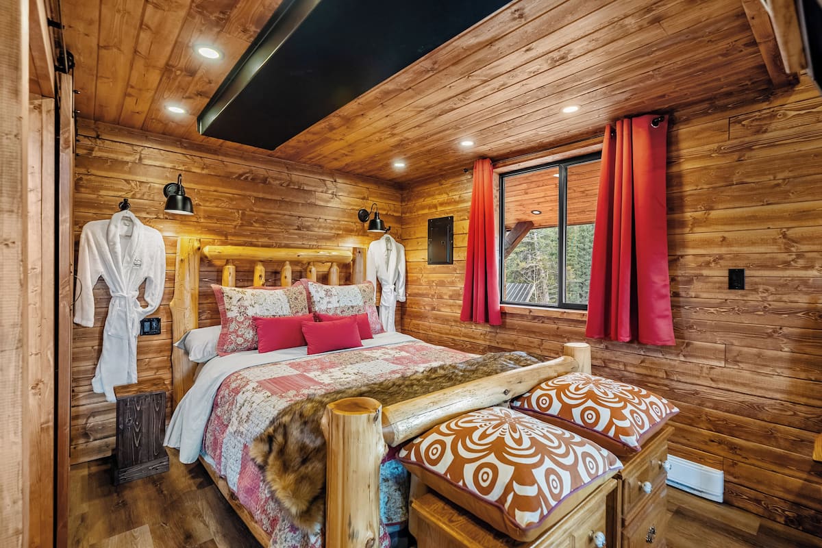 Un dormitori de cabana de fusta amb llit, almoadines, una taula de nit i una finestra amb cortines.