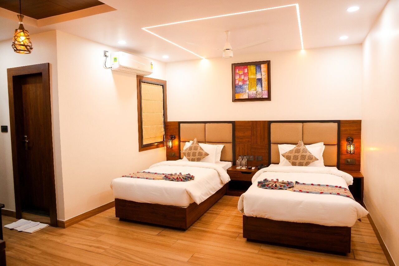 A hotel room with two beds, wooden headboards, a ceiling light fixture, a wall-mounted air conditioner, and a colorful artwork above the beds.