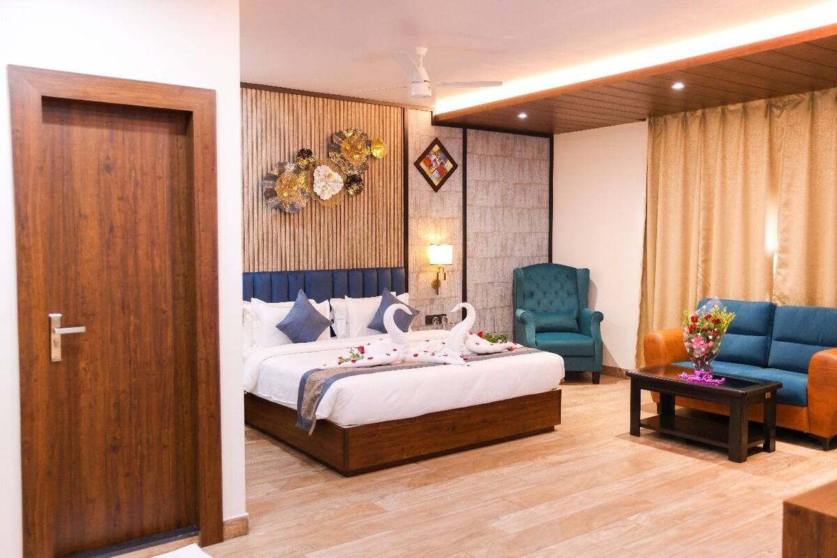 A hotel room with a wooden door, a bed with white bedding and decorative swans, a teal armchair, a blue sofa, and a small table with a vase of flowers.