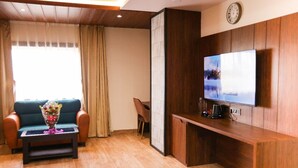 A living room with a sofa, a coffee table with a bouquet, a TV on a wooden stand, and a clock on the wall. - River sidde inn (Golmuri)